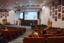 Widok dużej sali wykładowej podczas konferencji; uczestnicy siedzą w rzędach audytorium, a na scenie prelegenci prowadzą prezentację wyświetlaną na dwóch dużych ekranach. Na ścianach widoczne są portrety, a po obu stronach sceny stoją roll‑upy wydarzenia.