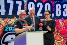 Trzy osoby stoją przy mównicy podczas wydarzenia charytatywnego. Dwie z nich trzymają mikrofony i notatki, a w tle widać duży kolorowy ekran z widocznymi cyframi i grafiką.”