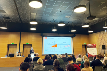 Publiczność siedzi w rzędach w sali konferencyjnej. Na scenie trzy osoby siedzą za stołem, a jedna stoi przy mównicy. Za mównicą znajdują się dwa duże ekrany z wykresami w kolorach pomarańczowych i niebieskich. W tle widać drewniane ściany i ciemny sufit.