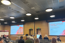 Sala konferencyjna z drewnianymi ścianami i dużymi wiszącymi lampami. Publiczność siedzi w kilku rzędach. Na scenie osoba stojąca przy mównicy prowadzi prezentację. Obok wyświetlone są dwa ekrany ze slajdami o nefrologii.