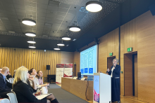 Sala konferencyjna z drewnianymi ścianami i ciemnym sufitem. Po lewej stronie siedzi kilkanaście osób na krzesłach. Po prawej stronie osoba prowadząca stoi przy białej mównicy i mówi do publiczności. Za prowadzącą znajduje się duży ekran z jasnym slajdem prezentacji.