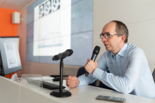 Osoba przy biurku prowadzi prezentację, trzymając mikrofon; obok niej znajduje się monitor z wyświetlonym slajdem oraz dodatkowy mikrofon na statywie.