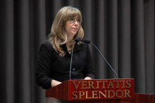 Osoba stojąca za mównicą z napisem „Veritatis Splendor”. W tle ciemna zasłona i dwa mikrofony ustawione na mównicy.