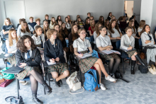 Grupa uczniów siedzi w sali podczas zajęć lub wykładu; wiele osób robi notatki, a przed pierwszym rzędem widoczne są plecaki i torby