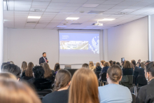 W sali wykładowej stoi osoba w garniturze i mówi do mikrofonu. Przed osobą siedzi grupa słuchaczy. 