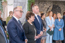 W jasnym wnętrzu stoi grupa kilkunastu osób ustawionych w dwóch rzędach. Na pierwszym planie trzy osoby stoją blisko siebie; jedna z nich trzyma tablet. Wszyscy mają formalne stroje. W tle widać osoby stojące pod ścianą z kamienną dekoracją oraz rośliny zwisające z góry.