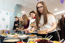 Osoby stoją przy stole z różnorodnymi składnikami do samodzielnego komponowania posiłków. Na stole znajdują się miski z warzywami, sałatkami i dodatkami, a uczestnicy nakładają jedzenie do papierowych misek.