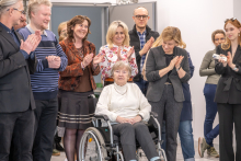 Na zdjęciu widać grupę kilkunastu osób stojących w pomieszczeniu z jasnym sufitem i ścianą wyłożoną kamiennymi elementami. Na pierwszym planie pośrodku znajduje się osoba siedząca na wózku, ubrana w jasną bluzę i ciemne spodnie. Wokół stoją osoby w różnych strojach.