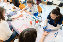 Osoby siedzą przy stolikach podczas akcji profilaktycznej. Jedna z osób ma mierzone ciśnienie krwi, a innej wykonywany jest pomiar glukozy glukometrem. Na stole leżą materiały informacyjne, rękawiczki i przyrządy medyczne.