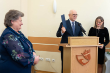 Mężczyzna stojący przy mównicy trzyma w dłoni dokument i przemawia podczas uroczystości; obok niego stoją dwie osoby.