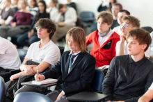 Grupa młodych osób siedzi w sali wykładowej; jedna z nich robi notatki w zeszycie, a pozostali słuchają prezentacji.