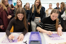 Dwie osoby siedzą przy stole i wykonują nadruki na materiałowych torbach, używając fioletowej farby i wałków. Na stole leży tacka z farbą, a za nimi stoi grupa uczestników obserwujących pracę.