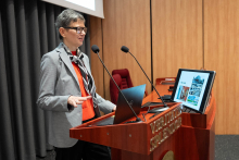 Osoba stojąca przy mównicy z laptopem i dwoma mikrofonami. 