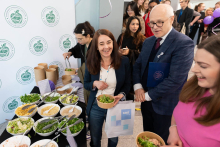 Osoby stoją przy stole z różnymi składnikami do przygotowania sałatek. Na stole ustawione są miski z warzywami, zieleniną i dodatkami, a w tle widoczna jest ścianka z logo instytutu. Uczestnicy wydarzenia nakładają produkty do papierowych misek.