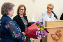 Podczas uroczystości jedna z osób wręcza bukiet kwiatów, a inna stoi przy mównicy z mikrofonem