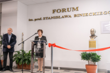 W jasnym holu stoją dwie osoby przy ścianie z dużym napisem ‘Forum im. prof. Stanisława Binieckiego’. Jedna osoba stoi za przezroczystą mównicą z mikrofonem, ubrana w ciemny strój. Druga osoba stoi z lewej strony, obok dużych zielonych roślin w donicach. Przed nimi rozciągnięta jest czerwona wstęga przymocowana do dwóch słupków. Po prawej stronie znajduje się popiersie ustawione na jasnym postumencie oraz roślina w doniczce.