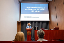 Osoba stojąca przy mównicy przed dużym ekranem z tytułem konferencji dotyczącym zdrowia jamy ustnej i opieki stomatologicznej nad osobami starszymi. 