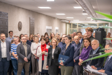 W jasnym korytarzu stoi duża grupa osób ustawionych blisko siebie. Wszyscy skierowani są w stronę mównicy widocznej po prawej stronie kadru. 