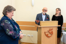 Podczas uroczystości mężczyzna stojący przy mównicy przemawia, wykonując gest ręką; obok niego stoi kobieta, a na pierwszym planie widoczna jest inna osoba słuchająca wystąpienia.