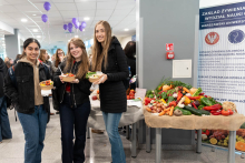 Trzy osoby stoją przy stole prezentującym warzywa, owoce i zdrowe produkty spożywcze. Każda z nich trzyma miskę z jedzeniem. Obok znajduje się roll‑up informacyjny Zakładu Żywienia Człowieka Warszawskiego Uniwersytetu Medycznego. W tle widać uczestników wydarzenia.