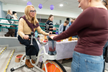 Osoba siedzi na rowerze stacjonarnym napędzając blender zamontowany z przodu urządzenia. Obok stoi druga osoba trzymająca pojemnik blendera. W tle widać stół z owocami i napojami oraz uczestników wydarzenia.