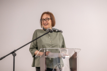 Na zdjęciu widać osobę stojącą za przezroczystą mównicą. 