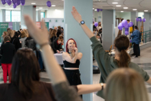 Osoby biorą udział w krótkiej sesji ćwiczeń lub rozciągania w przestrzeni wydarzenia. Na pierwszym planie widoczne są uniesione ręce uczestników, a w centralnej części osoba prowadząca wykonuje ćwiczenie. W tle znajdują się stoiska i inni uczestnicy