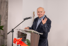 Na zdjęciu widać osobę stojącą za przezroczystą mównicą. Osoba ma na sobie ciemną marynarkę i koszulę w drobną kratkę. Jedna dłoń opiera się na mównicy, a druga jest uniesiona w geście. Przed mównicą ustawiony jest mikrofon na czarnym statywie. 