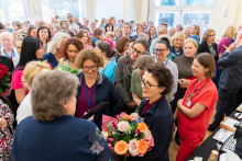 Wypełniona sala podczas uroczystości; w centrum grupa pracowników medycznych trzymających bukiet kwiatów, otoczona przez licznie zgromadzonych uczestników.