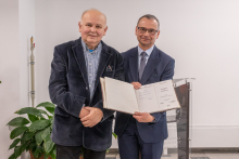 Na zdjęciu stoją obok siebie dwie osoby ubrane w eleganckie marynarki. Osoba po lewej ma ciemną marynarkę i koszulę w drobną kratkę, trzyma dłonie z przodu. Osoba po prawej ma granatowy garnitur i trzyma otwartą księgę, pokazując jej środek z widocznymi podpisami i tekstem. 