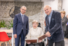 Na zdjęciu znajdują się trzy osoby w jasnym pomieszczeniu z dekoracyjną ścianą z kamiennych elementów. Pośrodku siedzi osoba na wózku, trzymająca dużą otwartą księgę. Po lewej stronie stoi osoba w eleganckim garniturze. Po prawej stronie stoi osoba w ciemnej marynarce, pochylona nad księgą, jakby pokazywała jej stronę. 