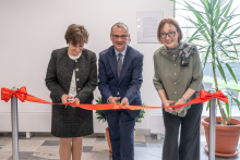 Na zdjęciu stoją trzy osoby ustawione w jednym rzędzie w jasnym pomieszczeniu. Każda z nich trzyma nożyczki i wspólnie przecina czerwoną wstęgę rozciągniętą między dwoma słupkami.  W tle widać białą ścianę, dużą roślinę doniczkową oraz tablicę z tekstem.