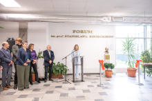 Jasne, przestronne wnętrze z napisem ‘FORUM im. prof. Stanisława Binieckiego’ na ścianie. Na środku stoi osoba przemawiająca przy przezroczystej mównicy z mikrofonem. Po lewej stronie grupuje się kilka osób stojących obok dużych roślin doniczkowych. Po prawej stronie widoczne jest popiersie ustawione na postumencie, ozdobione czerwoną wstążką. 