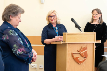 During the ceremony, a woman standing at a lectern delivers a speech, while two other people stand to the side listening.