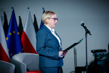 An audience seated in rows of red auditorium chairs. Women and men are looking toward the stage, and some people are holding documents or notebooks on their laps.