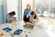 Several participants work at a table practicing medical procedures on manikins, while one person stands nearby observing.