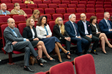 A group of attendees seated in the front rows of red chairs, facing the speaker on stage.