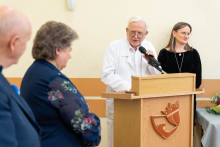 During the ceremony, a doctor in a white coat speaks at a lectern while holding a microphone; two other people are standing nearby, listening to the speech.