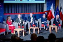 A panel discussion on stage. Five people are seated in armchairs arranged in a semicircle. The woman on the left is holding a microphone, and the other participants are listening. In front of the participants there are tables with glasses of water, and in the background there are flags and an institutional backdrop.
