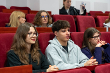 A group of younger event participants seated in an auditorium; some have their hands folded on the desk or resting on their laps.