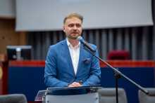 A man stands at a transparent lectern and speaks into a microphone, resting his hands on the surface of the lectern.
