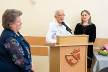 During the ceremony, a doctor in a white coat speaks at a lectern, while two other people are standing beside him.