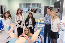 A group of participants stands around a gynecological training manikin placed on a hospital bed during a clinical skills workshop.