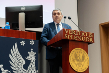A person standing at the “Veritatis Splendor” lectern, speaking into microphones; a computer screen and bottles of water are visible next to the podium.