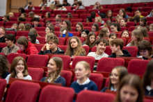  A group of young people sits in red auditorium seats; some talk to each other while others look toward the stage.