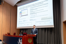 A person standing at a lectern, with a presentation slide displayed beside them showing text and a diagram related to medical documentation.
