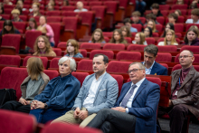 Several adult people sit in the front rows of red auditorium seats, facing the stage.