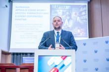 A speaker behind the lectern addressing the audience, with a large presentation slide behind showing the conference title in Polish and images related to pharmacy and crisis situations.