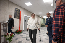 An elderly person using walking crutches walks through an indoor hall, accompanied by other attendees, with a commemorative plaque and flower arrangements visible in the background.