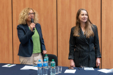Two people standing behind a conference table; one person is holding a microphone, and bottles of water, glasses, and documents are placed on the table.